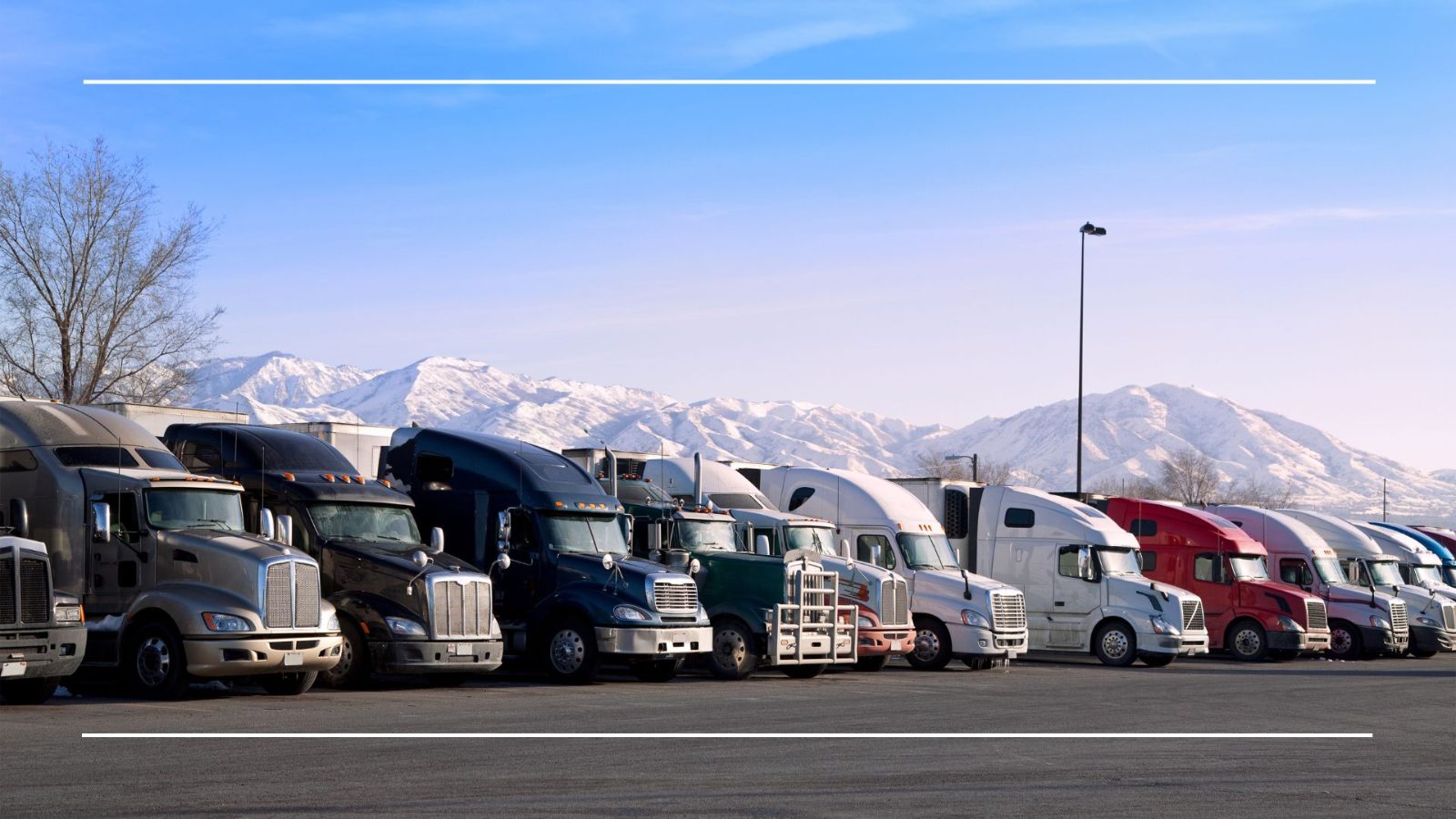 Row of parked semi trucks and trailers at a winter truck stop with snow-covered mountains behind